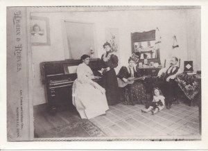 A photograph from the studio of Hearn & Reeves; Beulah Reeves at the piano,  her sister Augusta Reeves standing near, their brother  Walter Roger Reeves seated, his wife Mary Tennessee “Tennie” Harrison Reeves with guitar, and their son   Charles Walton Reeves on the floor, about age five, ca. 1897; courtesy Ruth Reeves Dill